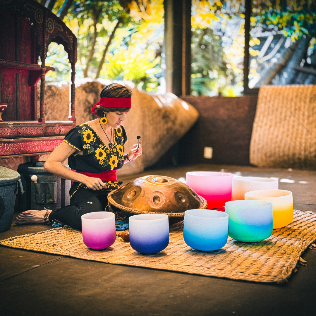 Person playing a handpan surrounded by colorful crystal singing bowls on a woven mat in a serene indoor setting.