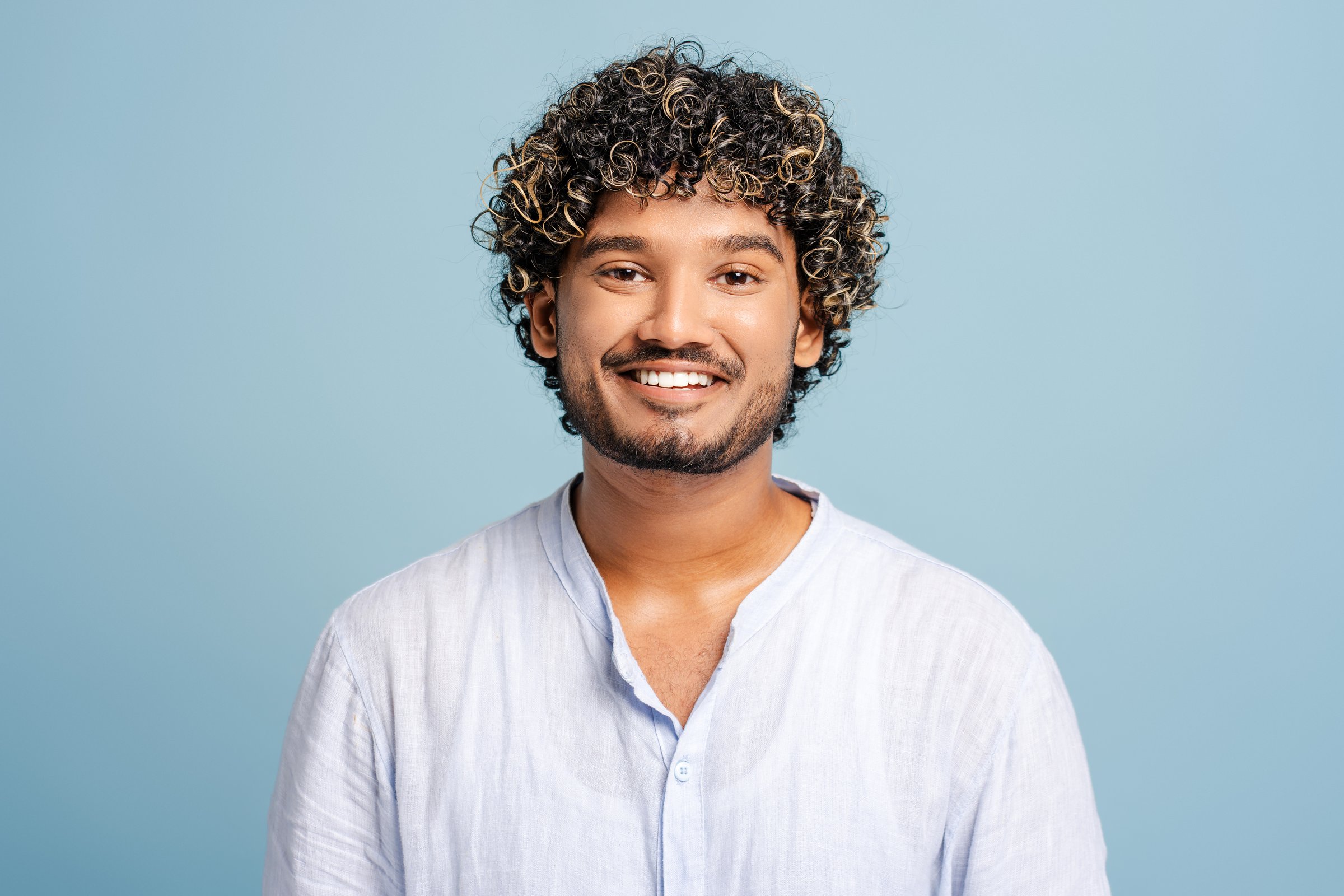 Smiling young man with curly hair on a blue background radiates happiness and warmth, capturing a carefree vibe