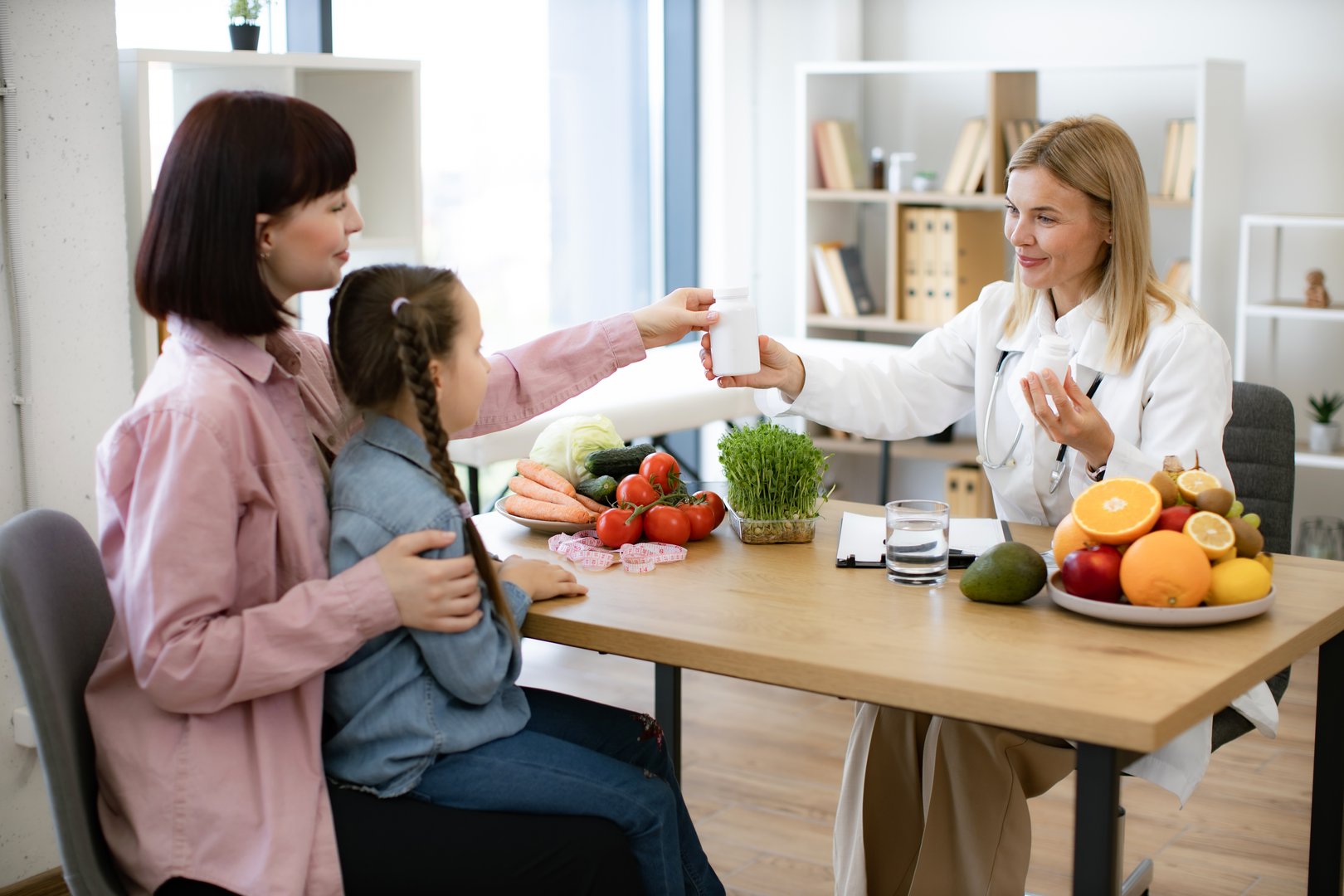 Mature pediatrician receives female patient in modern clinic. Happy pretty female nutritionist holding bottles with pills as nutrition supplements for little girl sitting on moms lap.