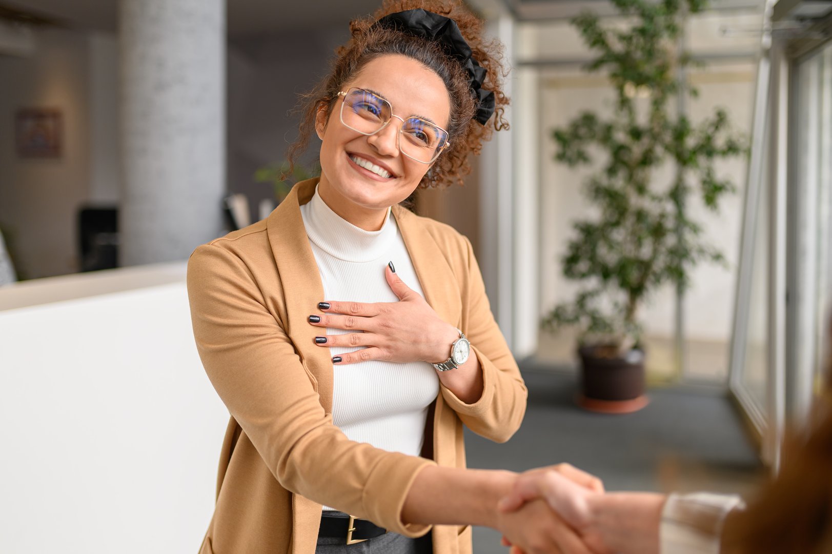 Smiling businesswoman in eyeglasses showing gratitude and shaking hands with female coworker in office