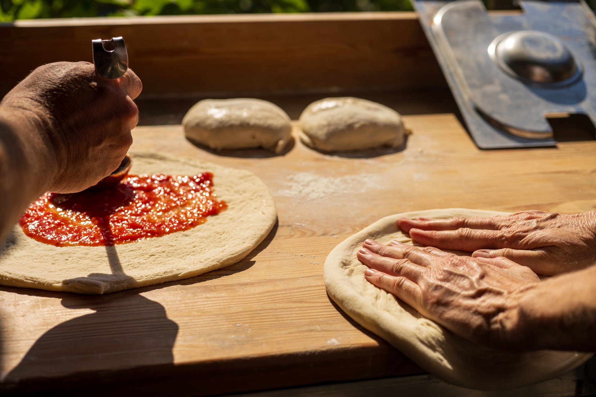 Close-up of two pairs of hands preparing pizzas on a rustic wooden counter. One hand presses the dough flat while the other spreads vibrant tomato sauce with a ladle. Soft natural light enhances the textures of the dough and sauce, evoking a handmade, farm-to-table culinary atmosphere.