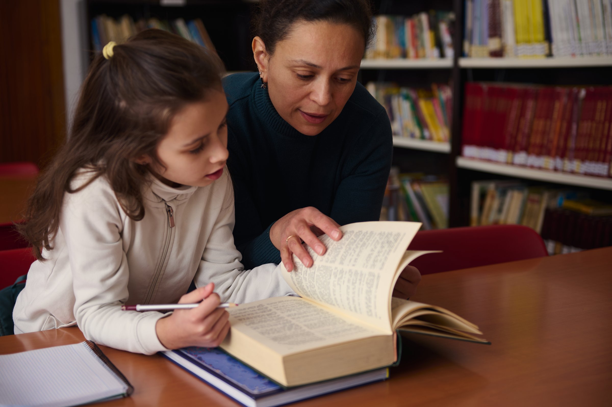 A woman guides a young girl through a book in a quiet library, fostering reading, learning, and focus in a collaborative educational moment.