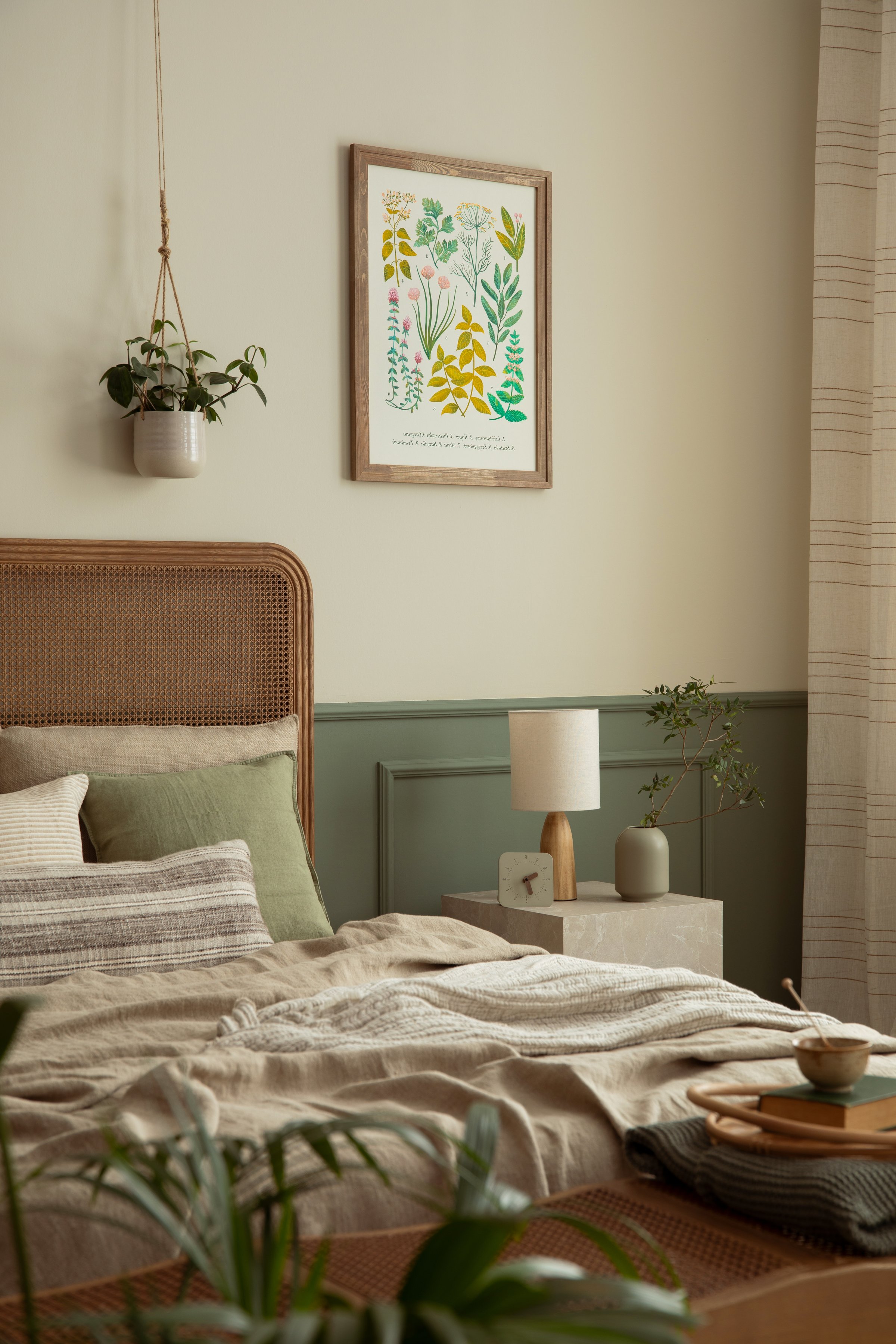 Cozy composition of bedroom interior with mock up poster frame, stylish bed, beige bedding, lamp, plants in pots, vase with leaves, wall with stucco and personal accessories. Home decor. Template.