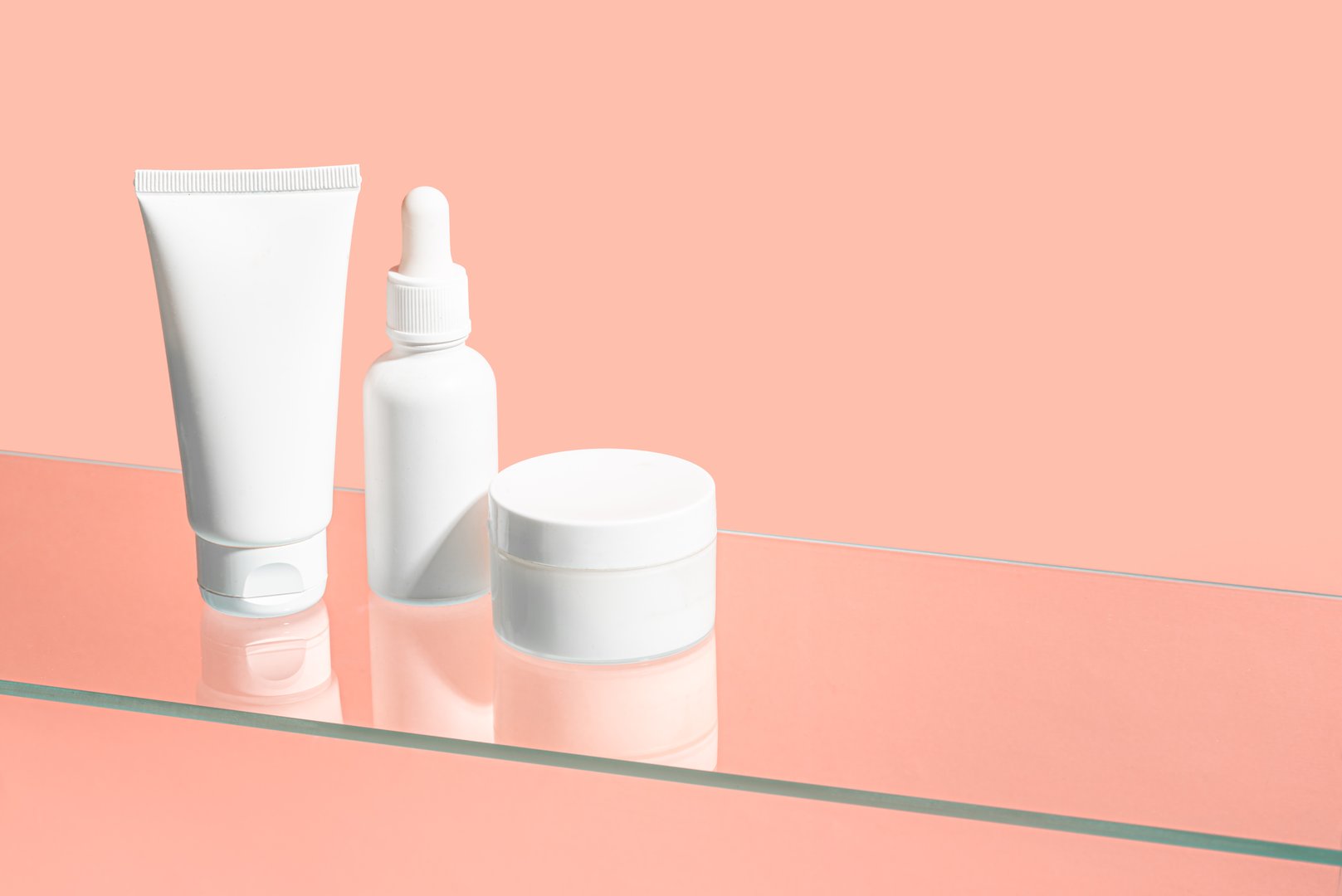 Cosmetic products containers on glass shelf and pink background, copy space. White blank face moisturizer cream jar, cleanser gel tube, face serum bottle branding, daily skin care routine.