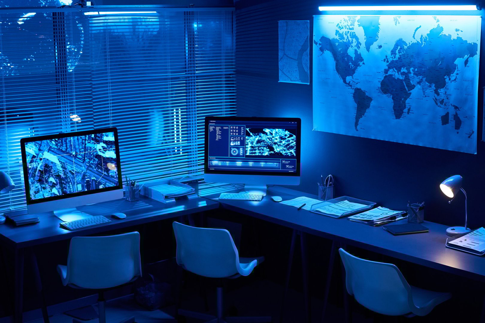 Desks with computer monitors with location data on screens standing along wall with map and window with venetian blinds