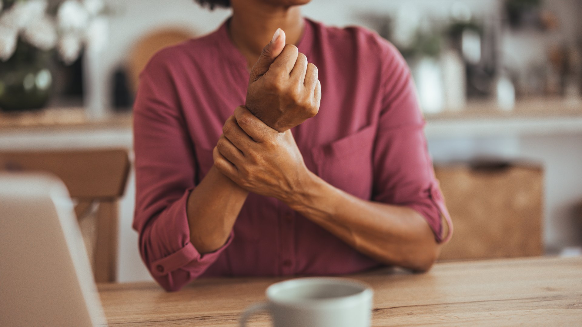 A woman in a pink shirt sits at a table, holding her wrist in discomfort, symbolizing wrist pain or arthritis in a home setting.