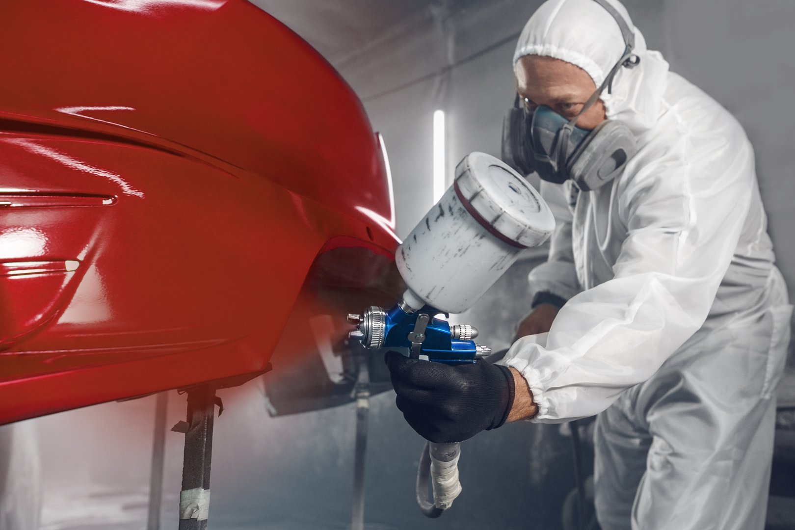 Professional car painter in chamber paints automobile bumper to red color. Man spraying finishing layer of paint to vehicle part.