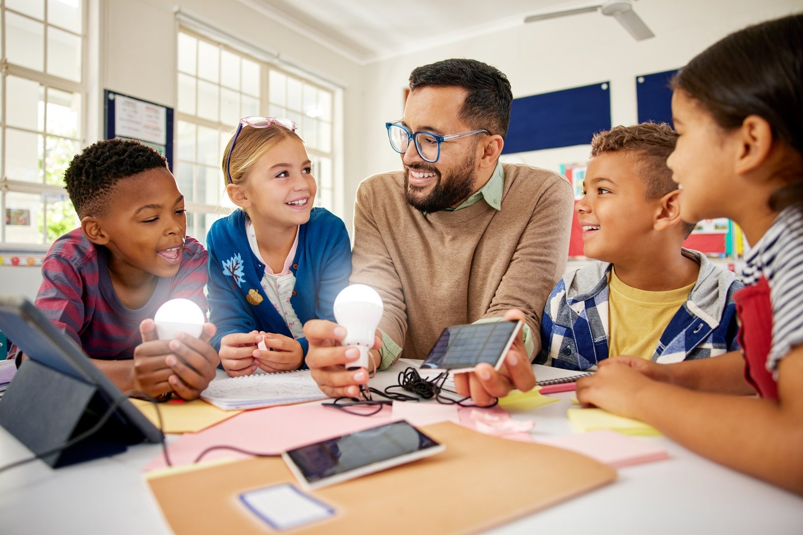 Happy male teacher with solar panel explaining pupil about solar energy with illuminated bulbs. Teacher in science classroom showing a solar panel to generate electricity. Smiling multiethnic primary children learning and experimenting about renewable energy sitting together around desks.