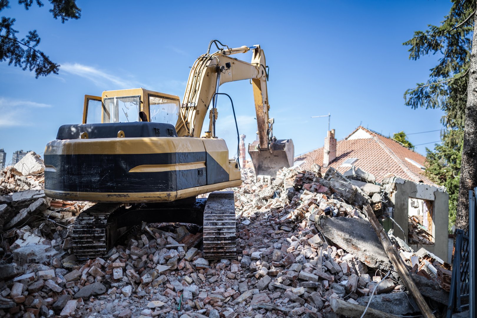 An excavator is actively clearing debris from a recently demolished structure, with remnants scattered across the site beneath a bright blue sky.