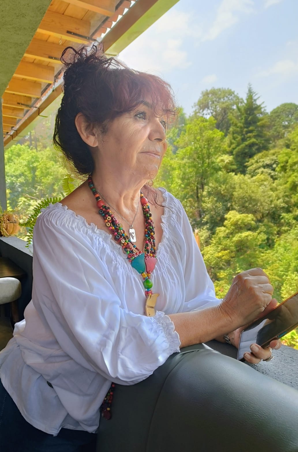 Elderly woman with red hair in a white blouse on a balcony, holding an object and gazing at a lush green landscape.