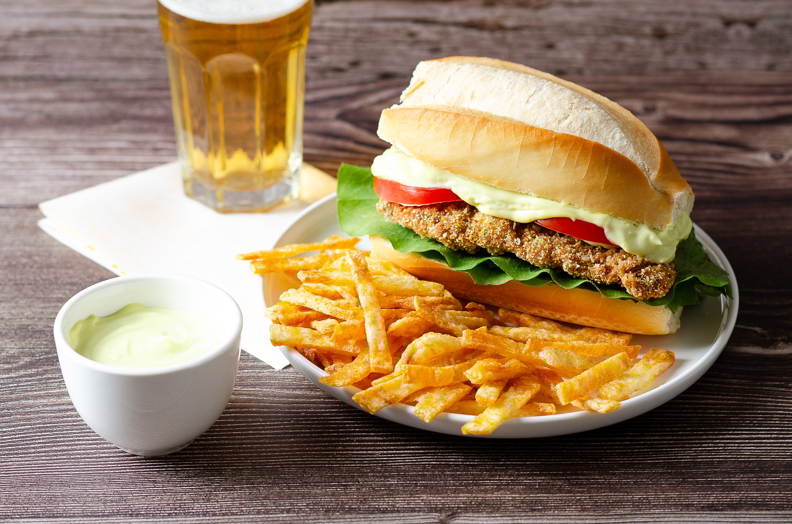 Milanesa sandwich with tomato slices, lettuce and mayonnaise and French fries in a white plate, a bowl with mayonnaise and a glass of beer on a wooden background.