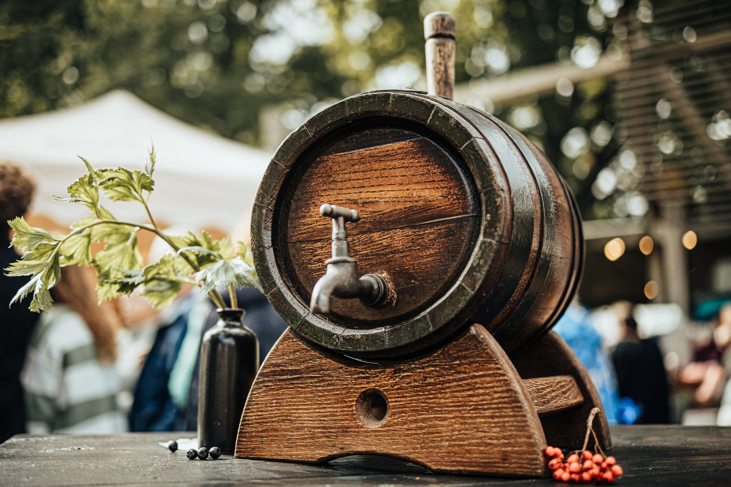 A rustic wooden barrel with a metal tap sits on a stand at an outdoor festival, decorated with greenery and berries, creating a traditional festive atmosphere