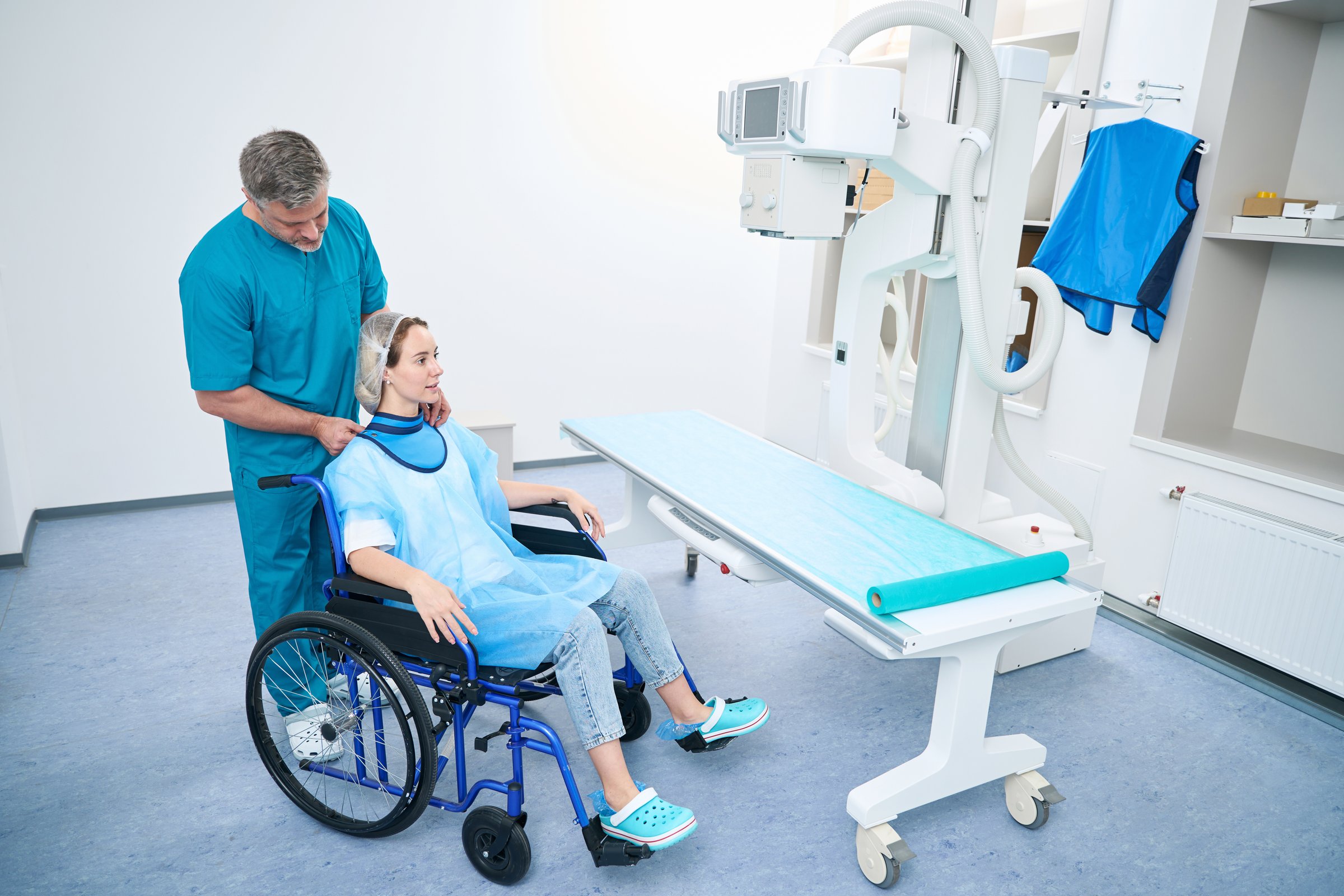 Attentive doctor in x-ray room putting lead protection on young woman in wheelchair