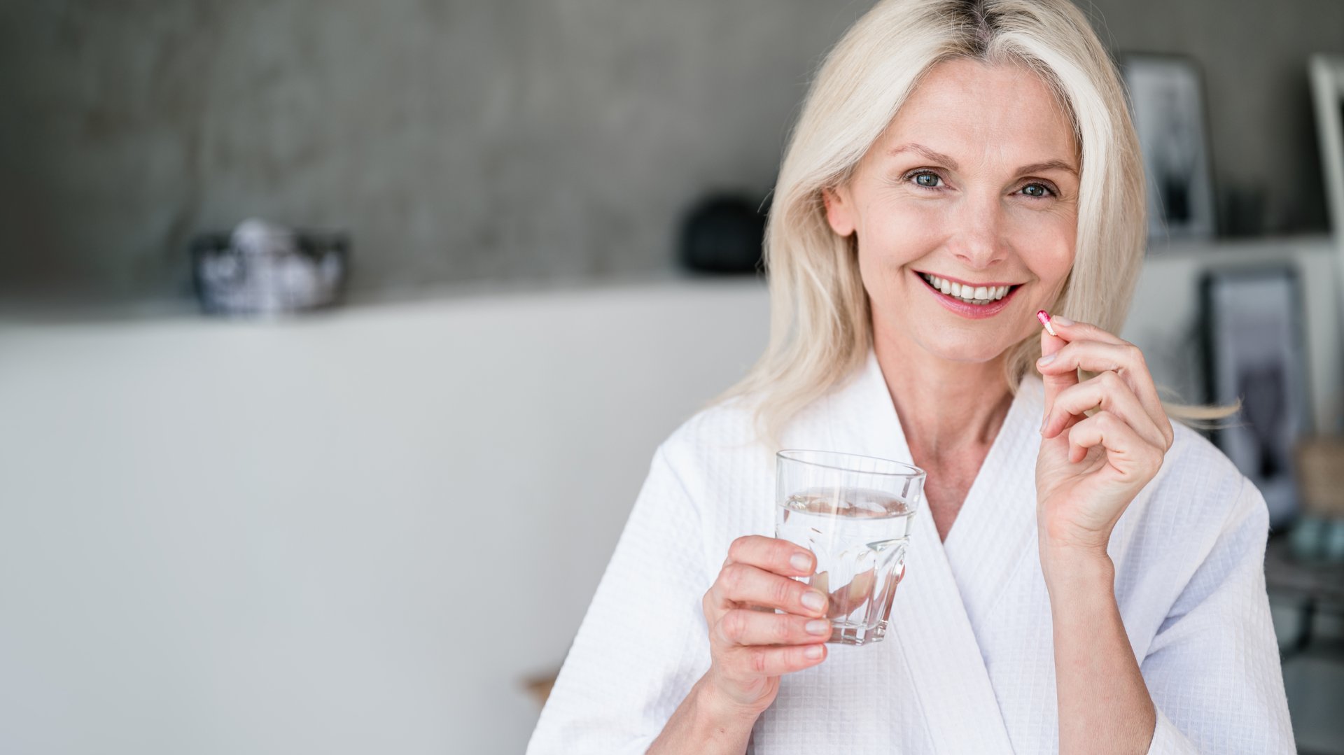 Web banner view of happy mature woman holding glass of still water, and vitamin pill with calcium and collagen. Mid age female feel good, smiling nice, taking dose of daily multivitamins at home