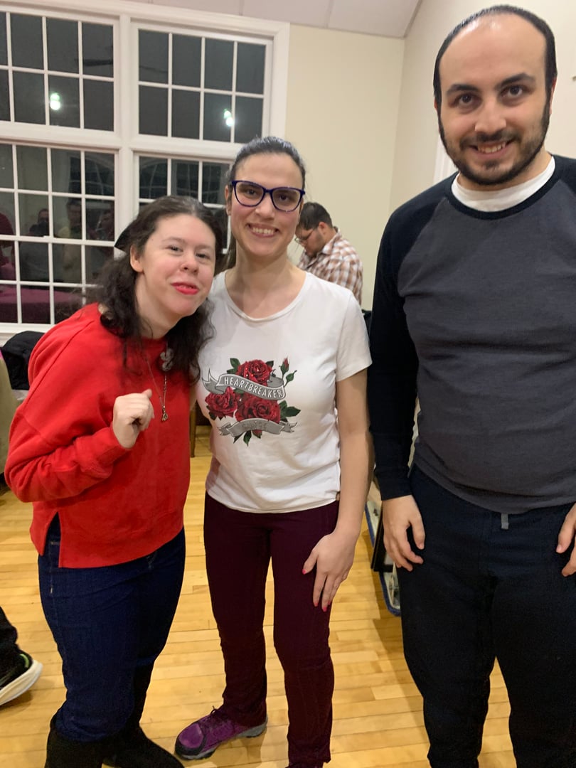 Three people smiling indoors, standing on a wooden floor with large windows in the background.