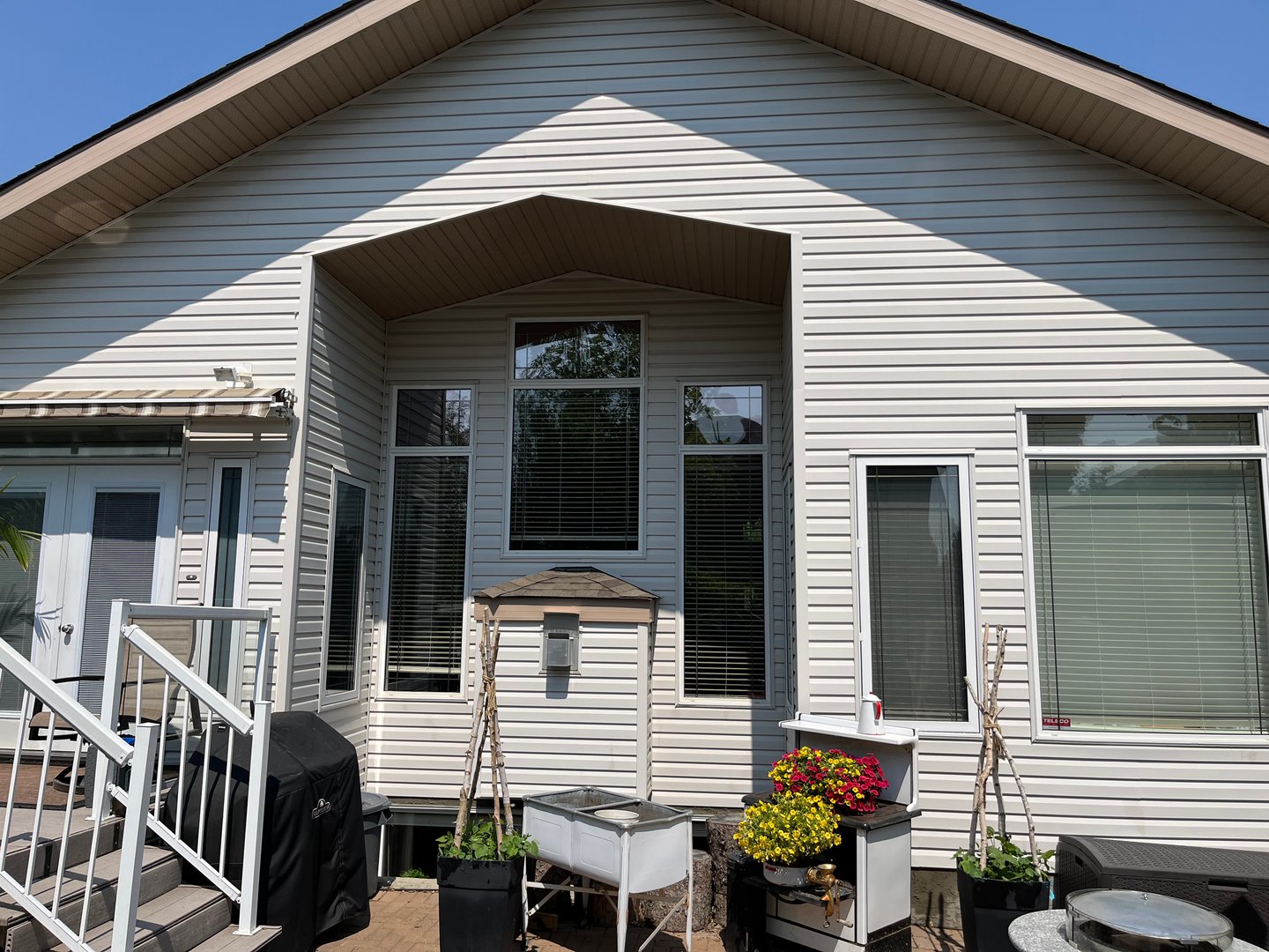 Backyard view of a house with white siding, several tall windows, and potted plants on a patio beside a barbecue grill.