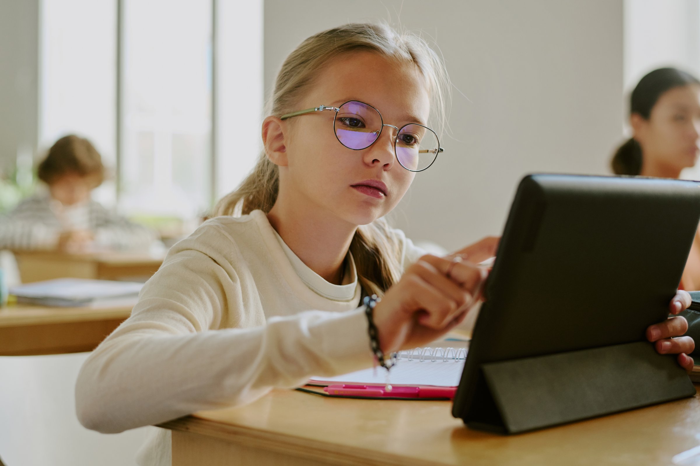 Young girl wearing glasses sitting at wooden desk using tablet in classroom surrounded by fellow students in bright learning environment