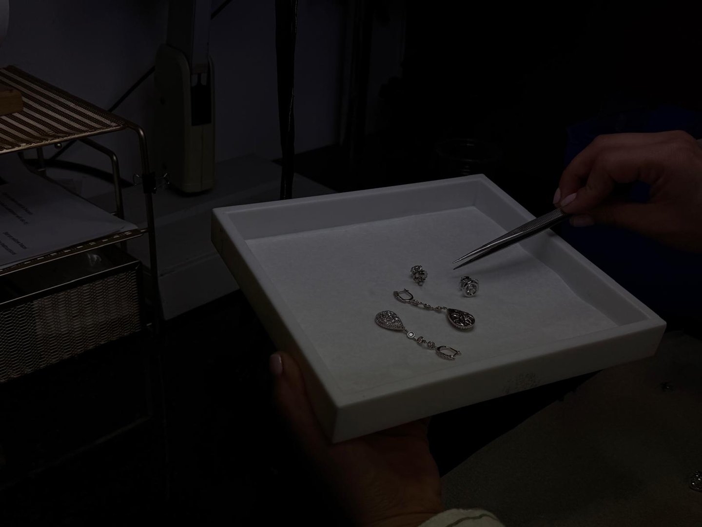 Person using tweezers to handle jewelry pieces on a tray in a dimly lit setting.