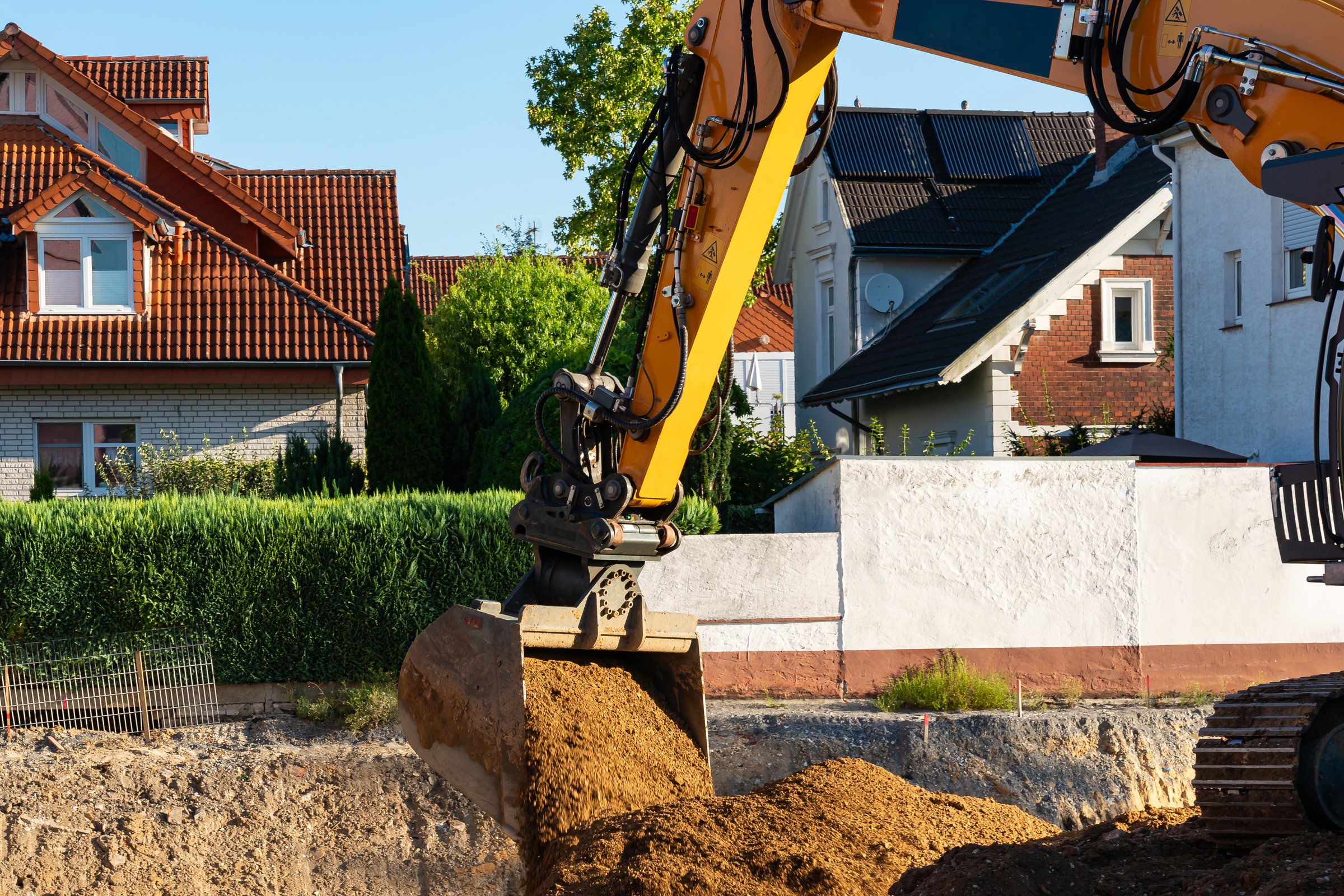A large excavator with a sturdy arm is digging into the ground, moving soil for construction work beside houses on a bright, sunny afternoon.
