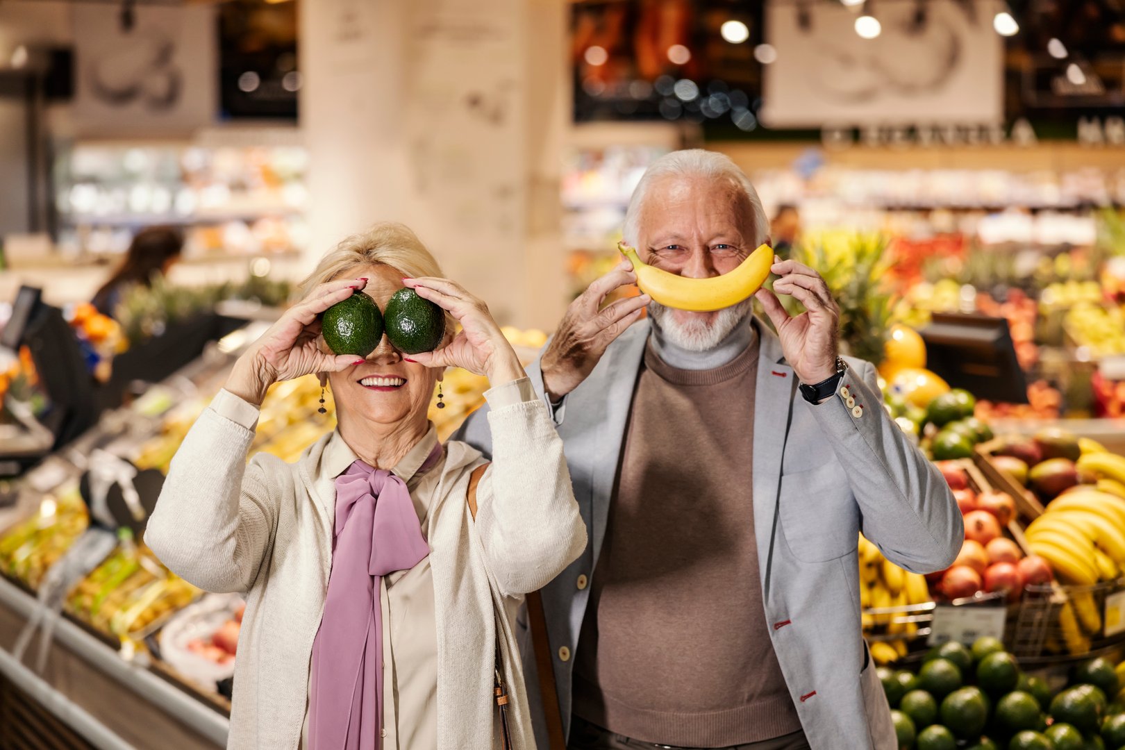 An old couple is playing silly faces with fruits at supermarket.