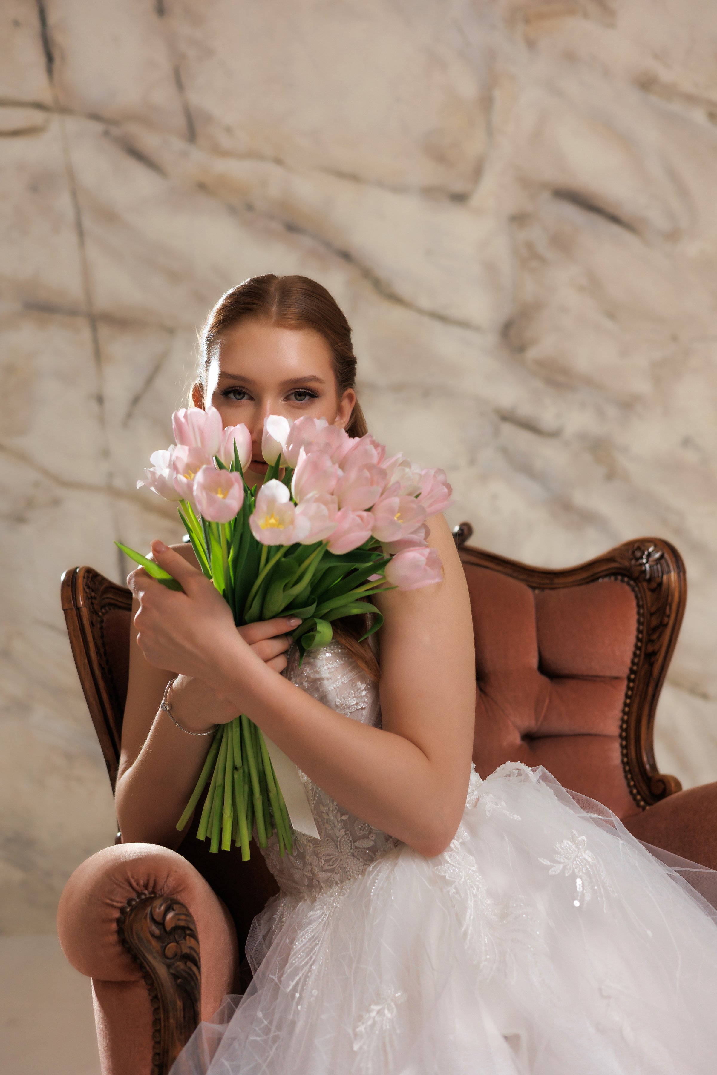Bridal portrait features a woman in a stunning wedding dress, playfully hiding her face behind a vibrant bouquet of pink tulips while relaxing on a vintage chair.