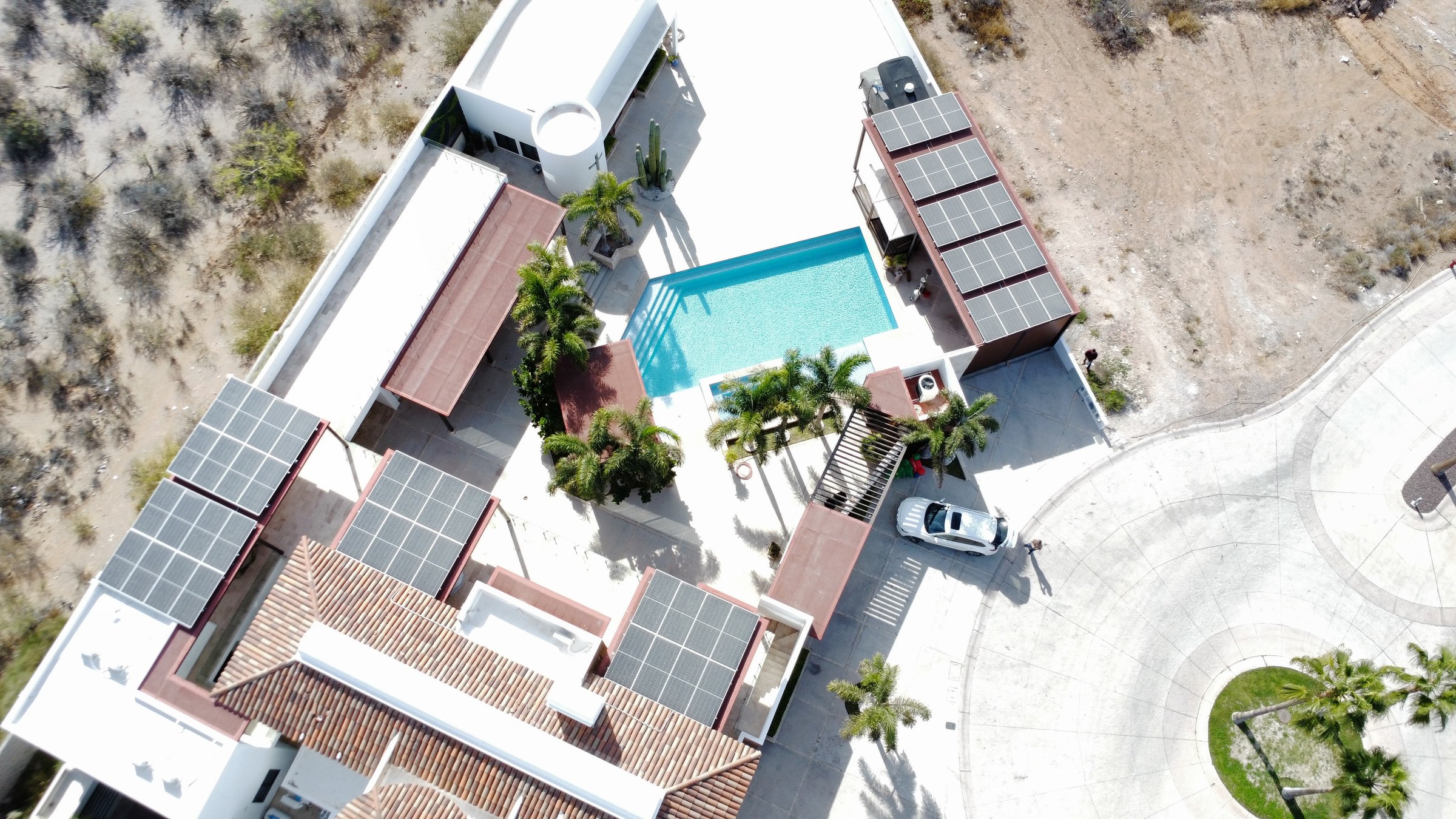 Aerial view of a house with solar panels and a swimming pool surrounded by palm trees and a driveway with a parked car.