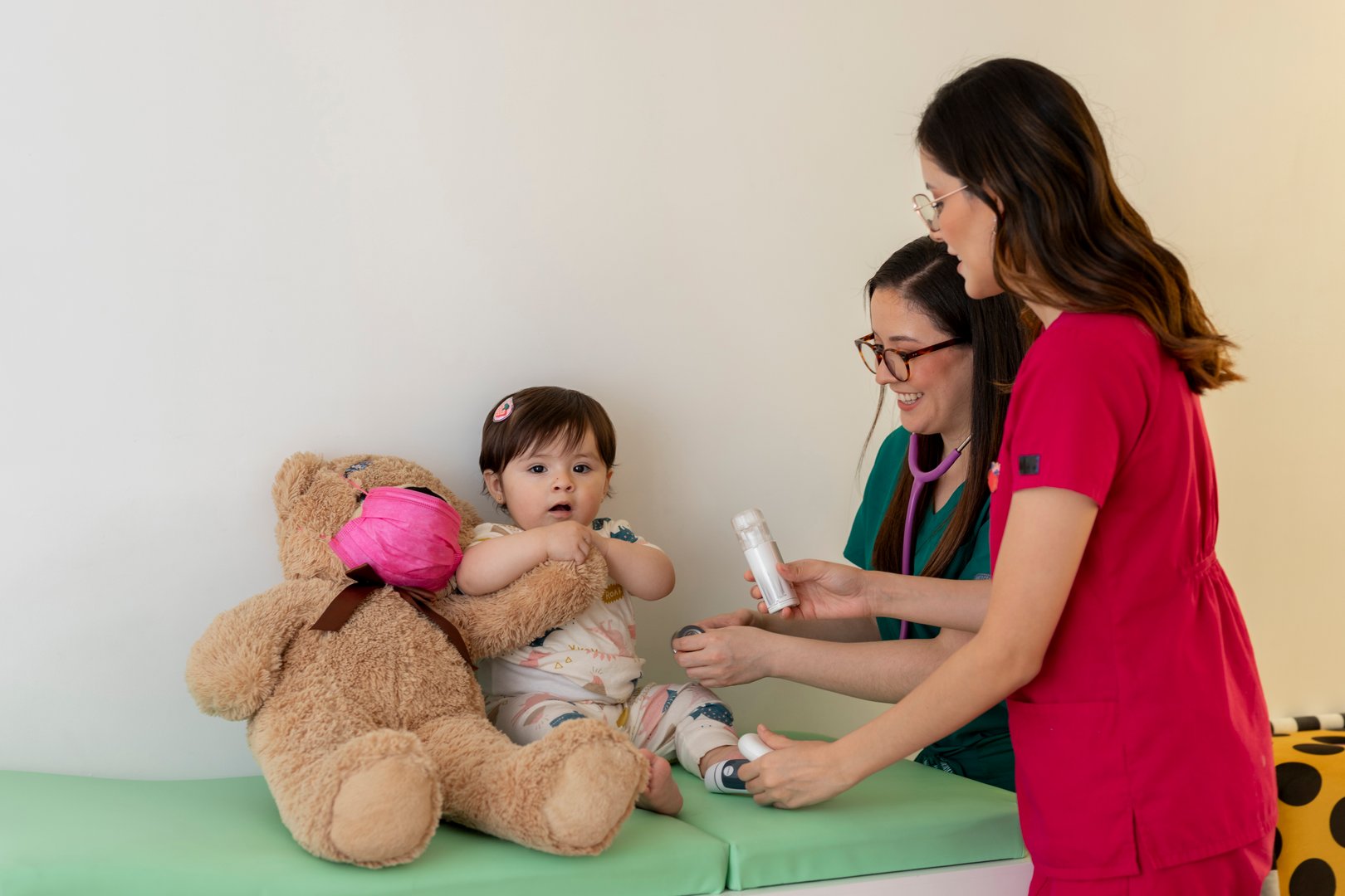 Two pediatricians are taking care of a baby girl sitting next to a teddy bear in a doctor's office