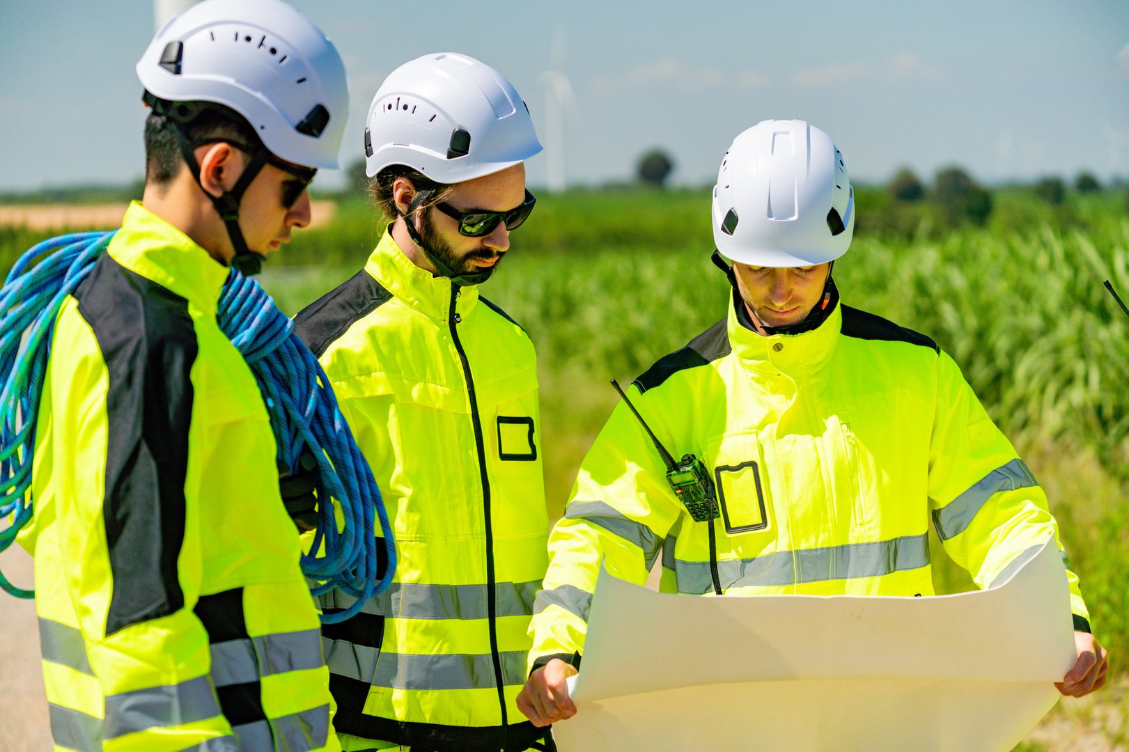 A group of three engineers in bright safety clothing studies construction plans. They are outdoors on a sunny day, surrounded by crops and clear blue skies.