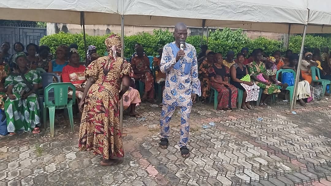A man in traditional attire speaks into a microphone under a canopy, with seated people in colorful clothes listening.