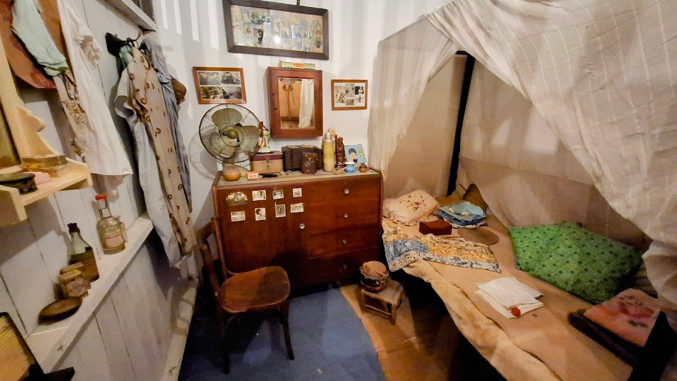 A small, cluttered room with a single bed, wooden desk, fan, and various personal items. Photos and clothes are on display.