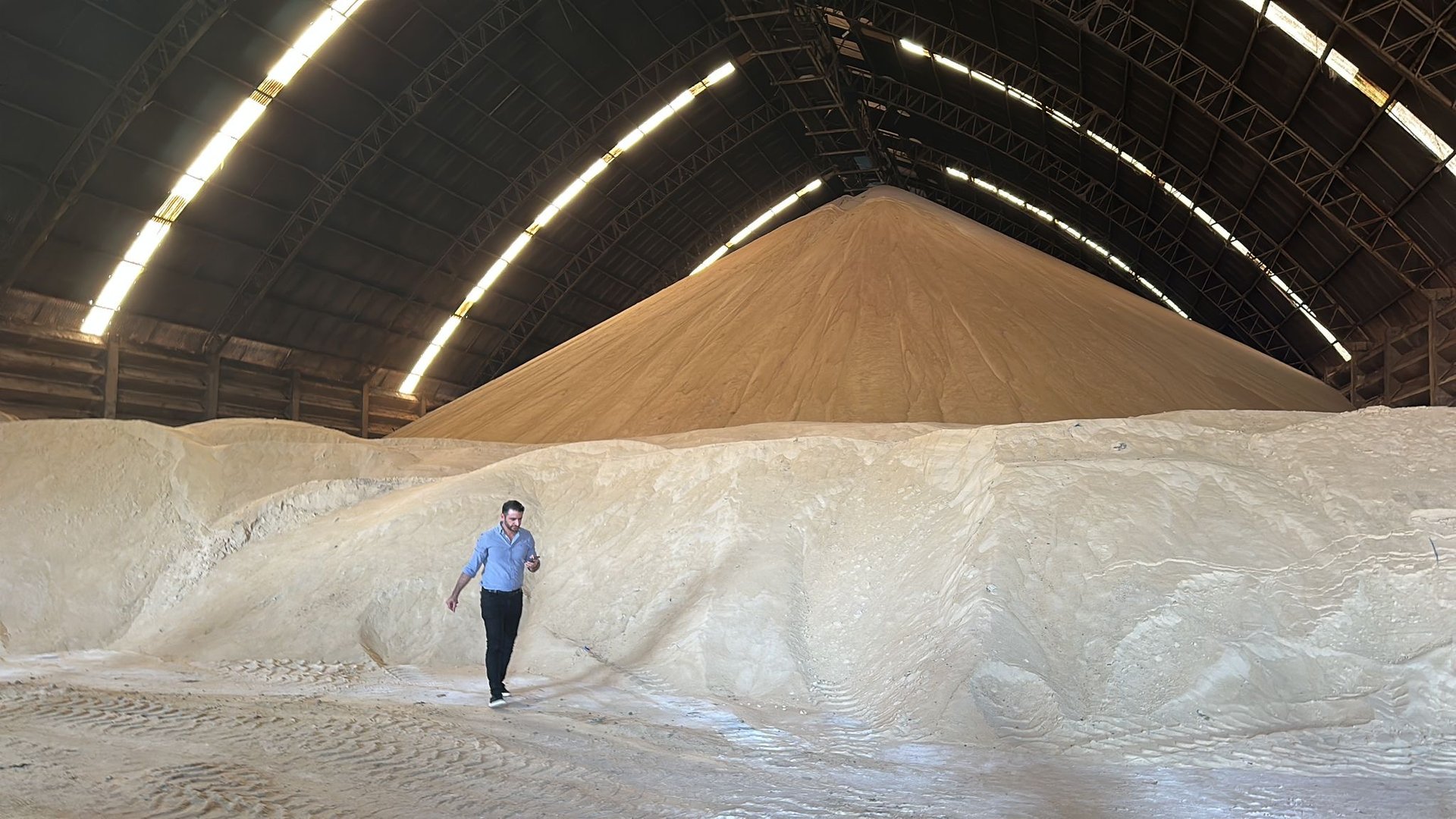 A person walking near a large pile of salt inside a well-lit industrial warehouse with a high, curved roof.