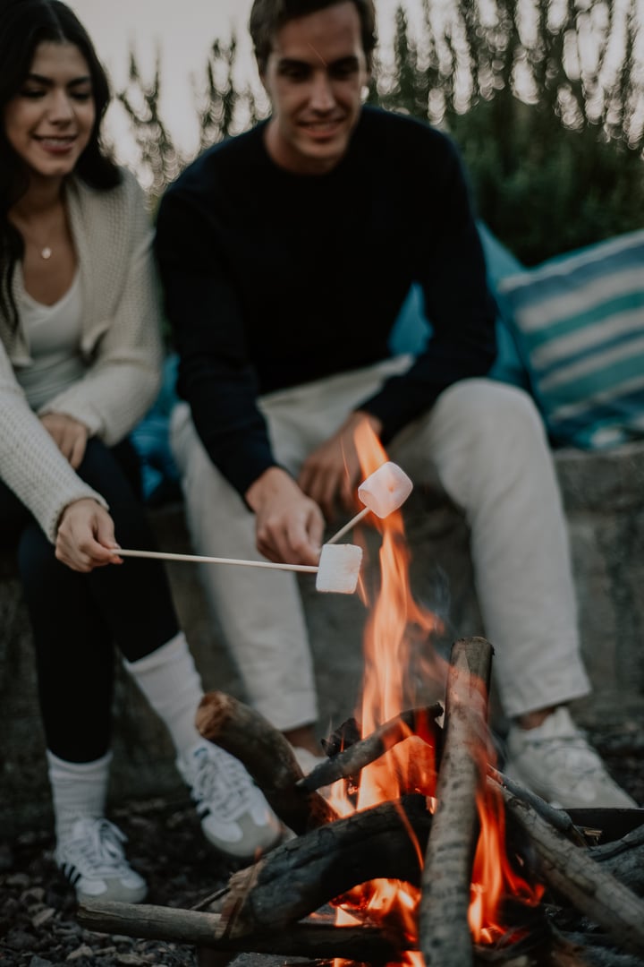 Two people roasting marshmallows over a campfire with wooden sticks, seated outdoors.