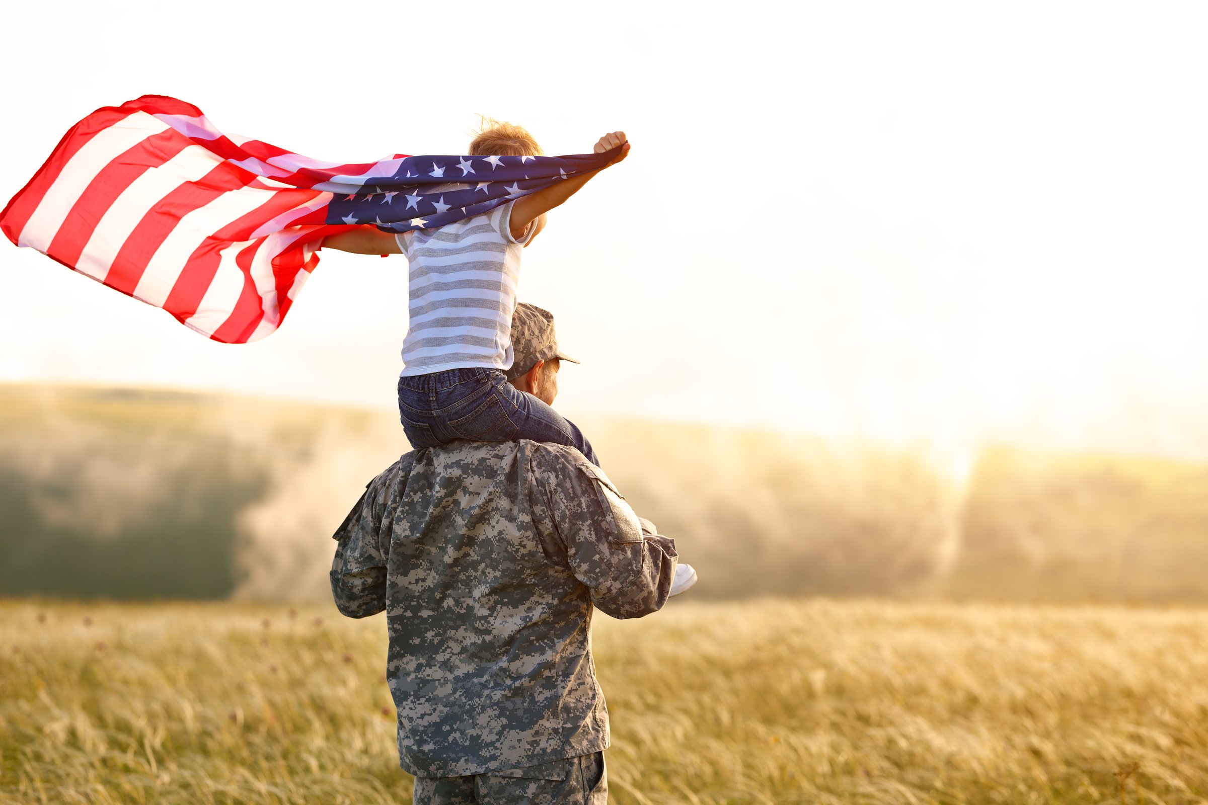 Rear view of military man father carrying happy little son with american flag on shoulders and enjoying amazing summer nature view on sunny day, happy male soldier dad reunited with son after US army
