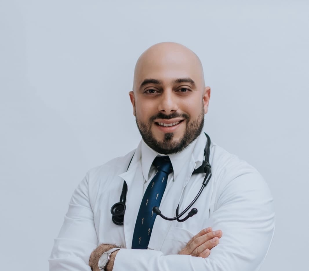 A smiling doctor with a bald head and beard, wearing a white coat and stethoscope, standing with arms crossed.