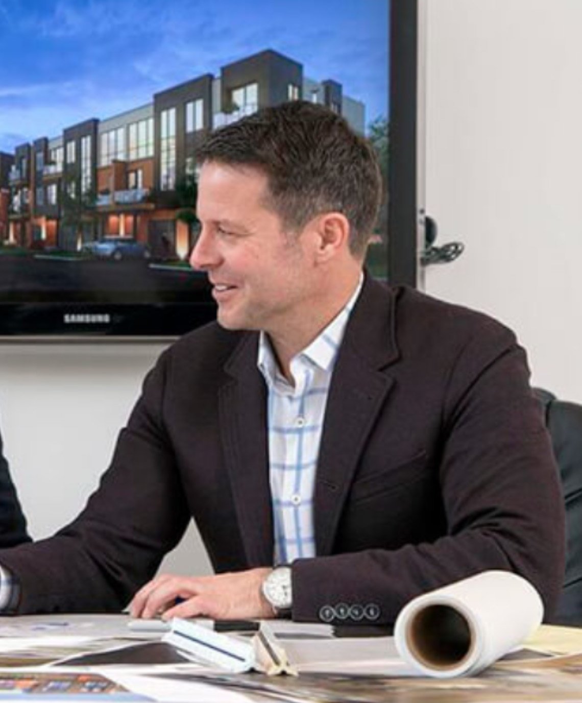 Man in a blazer and shirt sitting at a desk with architectural plans and a monitor displaying building designs.