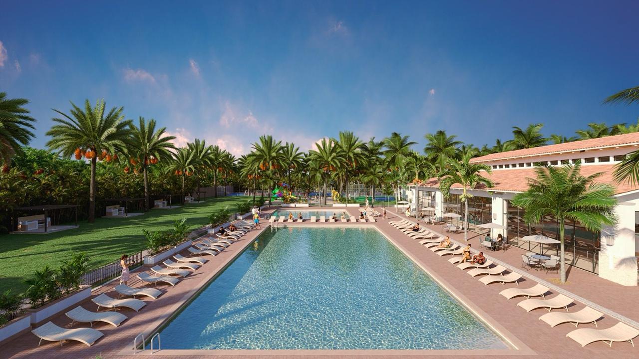 Resort pool surrounded by lounge chairs and palm trees, with a clear blue sky and guests relaxing in the sun.
