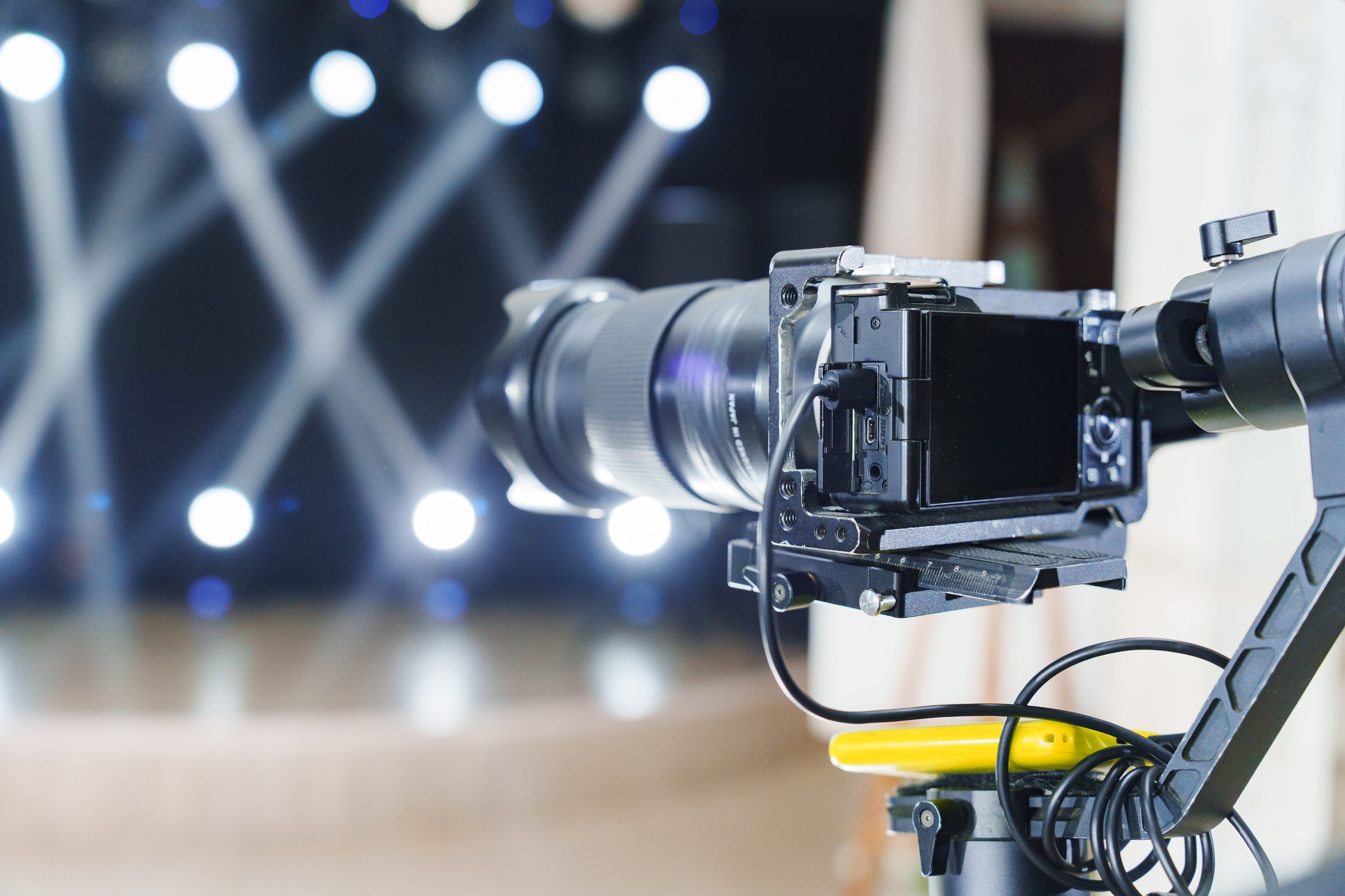 A camera is positioned on a tripod facing a stage. Bright lights are shining in the background, indicating a live performance or event is about to begin. The atmosphere is filled with anticipation.