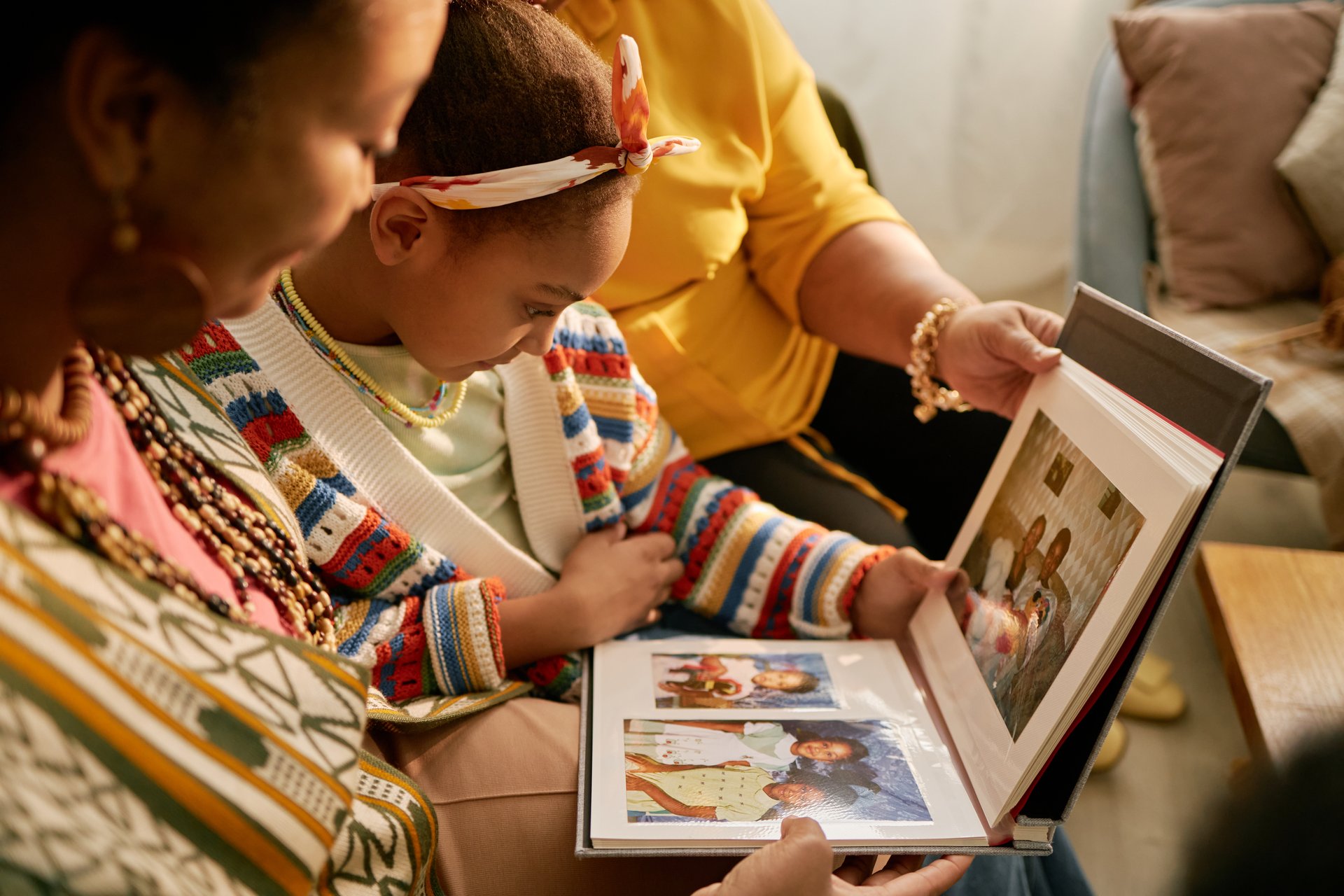 Black girl sitting with adults, enjoying family photo album. Group reminiscing about past moments and cherishing memories together in living room