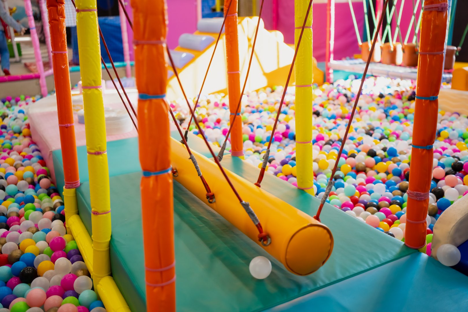 Colorful soft play balance bridge suspended above a ball pit inside an indoor playground, featuring padded posts, bright mats, and a fun obstacle designed for young children
