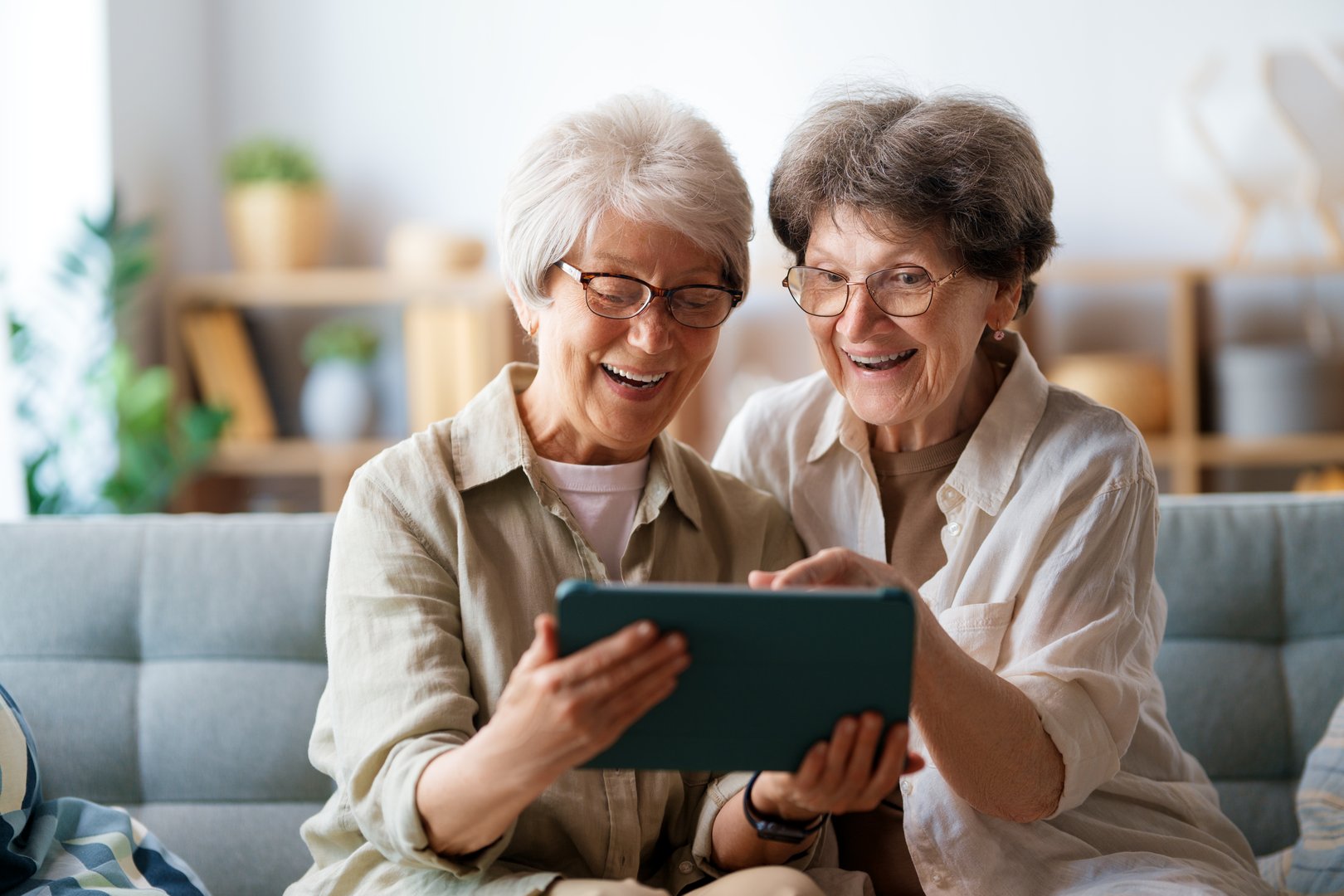 Happy senior women are using digital tablet while sitting together on the comfortable couch at home.