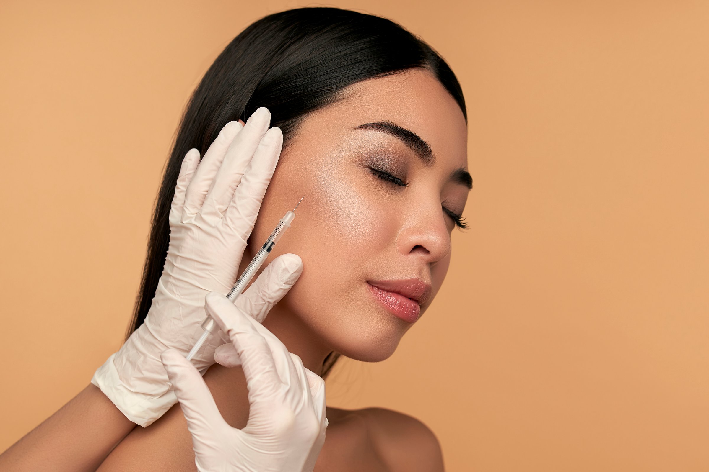 Young Asian woman with clean radiant skin gets botox injections for contour tightening, lip augmentation on a beige background. Spa care, facial skin care, beauty cosmetology.