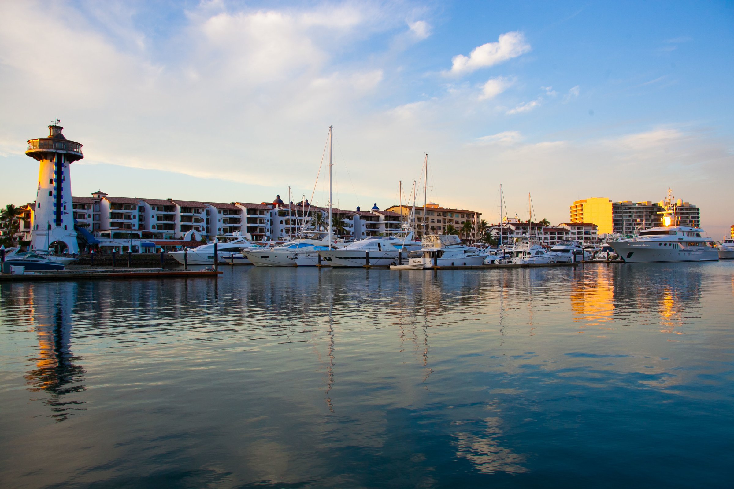 Marina at sunset with yachts docked and buildings in the background. Calm water reflects the boats and sky.