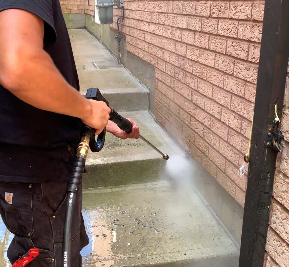 Person power washing outdoor concrete steps near a brick wall, removing dirt and debris with a high-pressure washer.