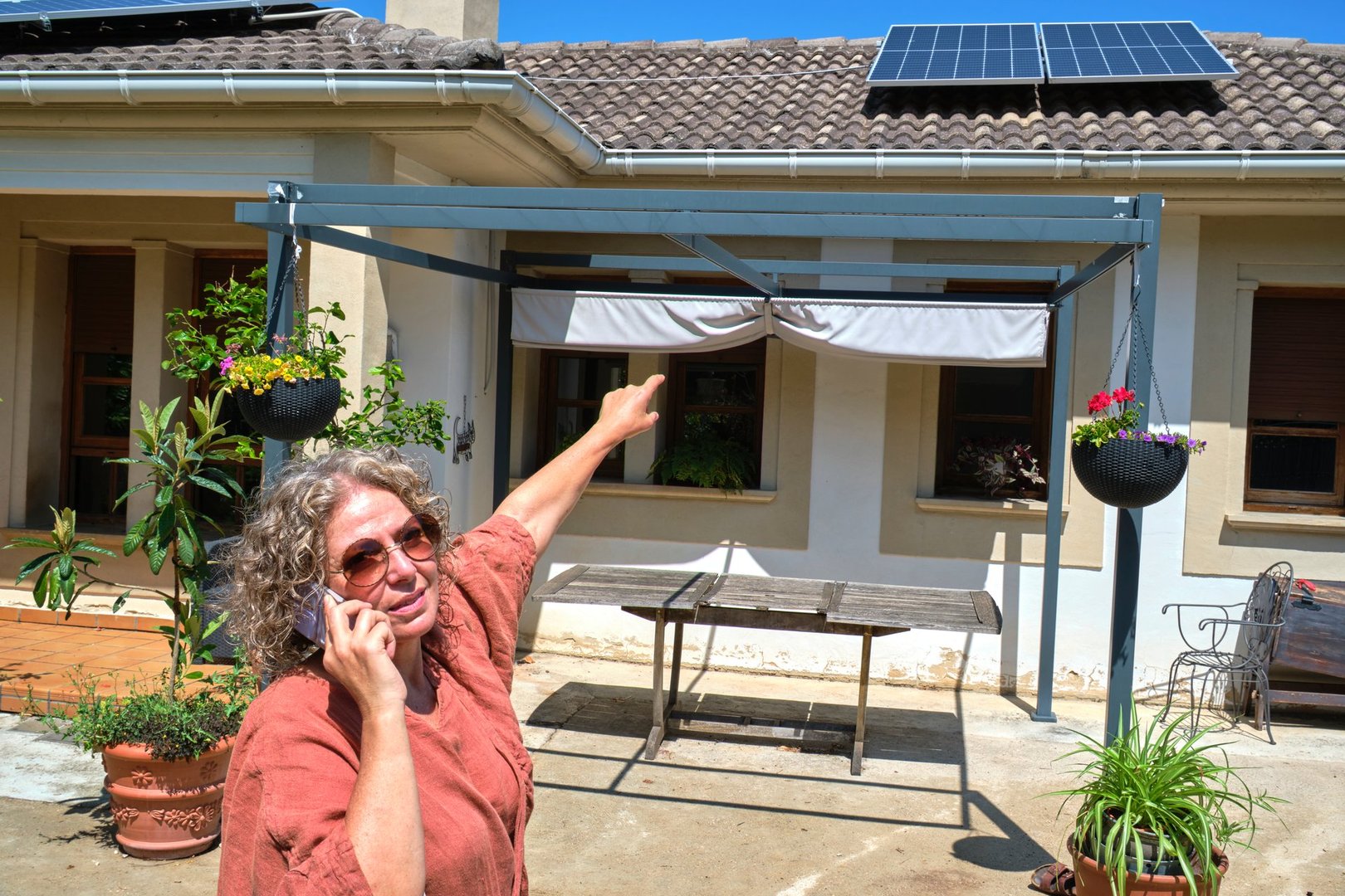 A woman wearing sunglasses is talking on the phone while pointing at solar panels on the roof in a sunny backyard. The setting includes potted plants and reflects a warm, eco-friendly atmosphere.