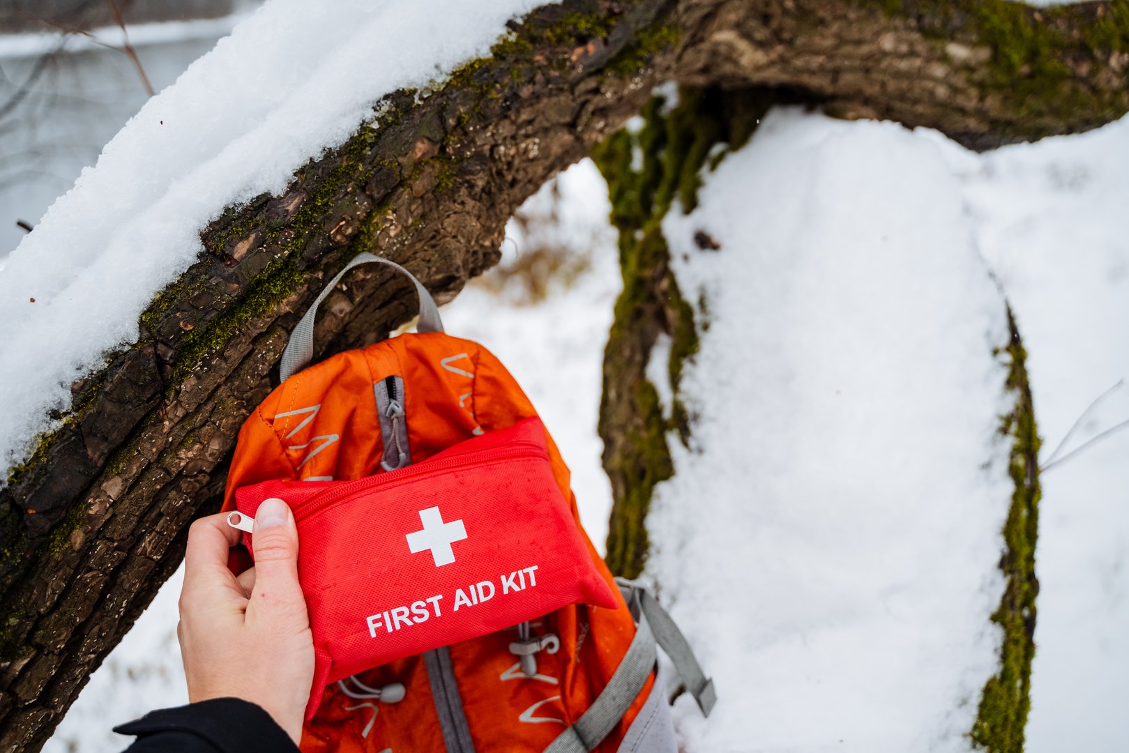 Person standing in snow holding first aid kit next to backpack