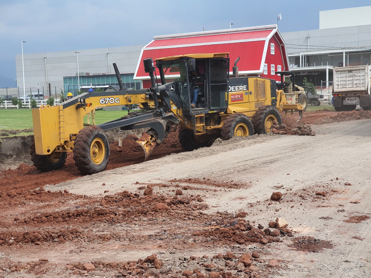 Yellow grader leveling red dirt on a construction site near a building with a red roof, under a cloudy sky.