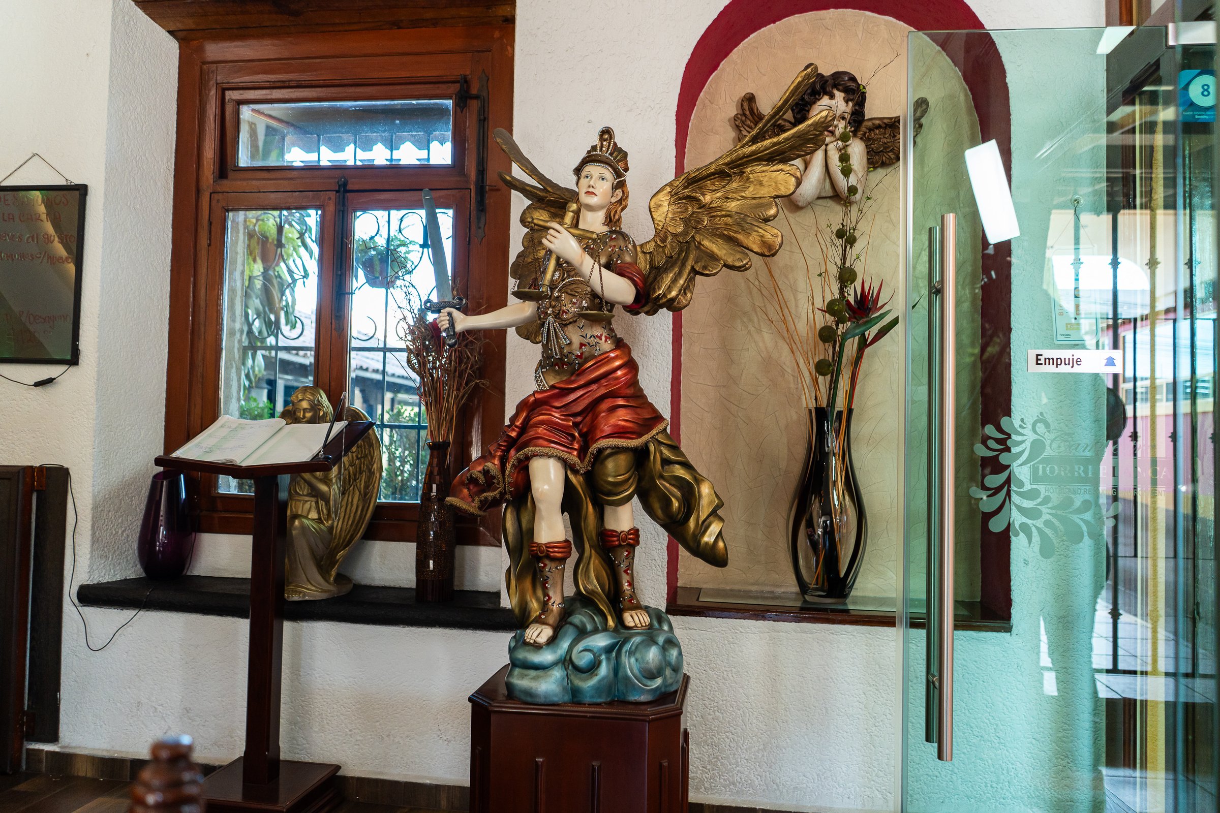 Angel statue with wings in decorative indoor setting, next to a window and glass door with floral elements.