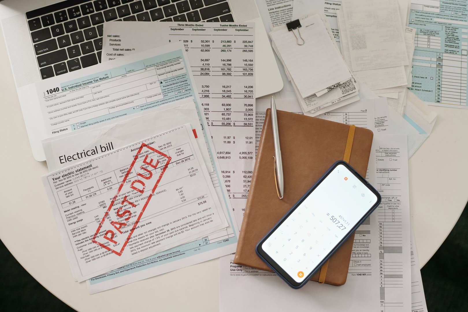 Overview of laptop keyboard, electrical bill with past due stamp, notebook in brown leather cover and smartphone with calculator on screen
