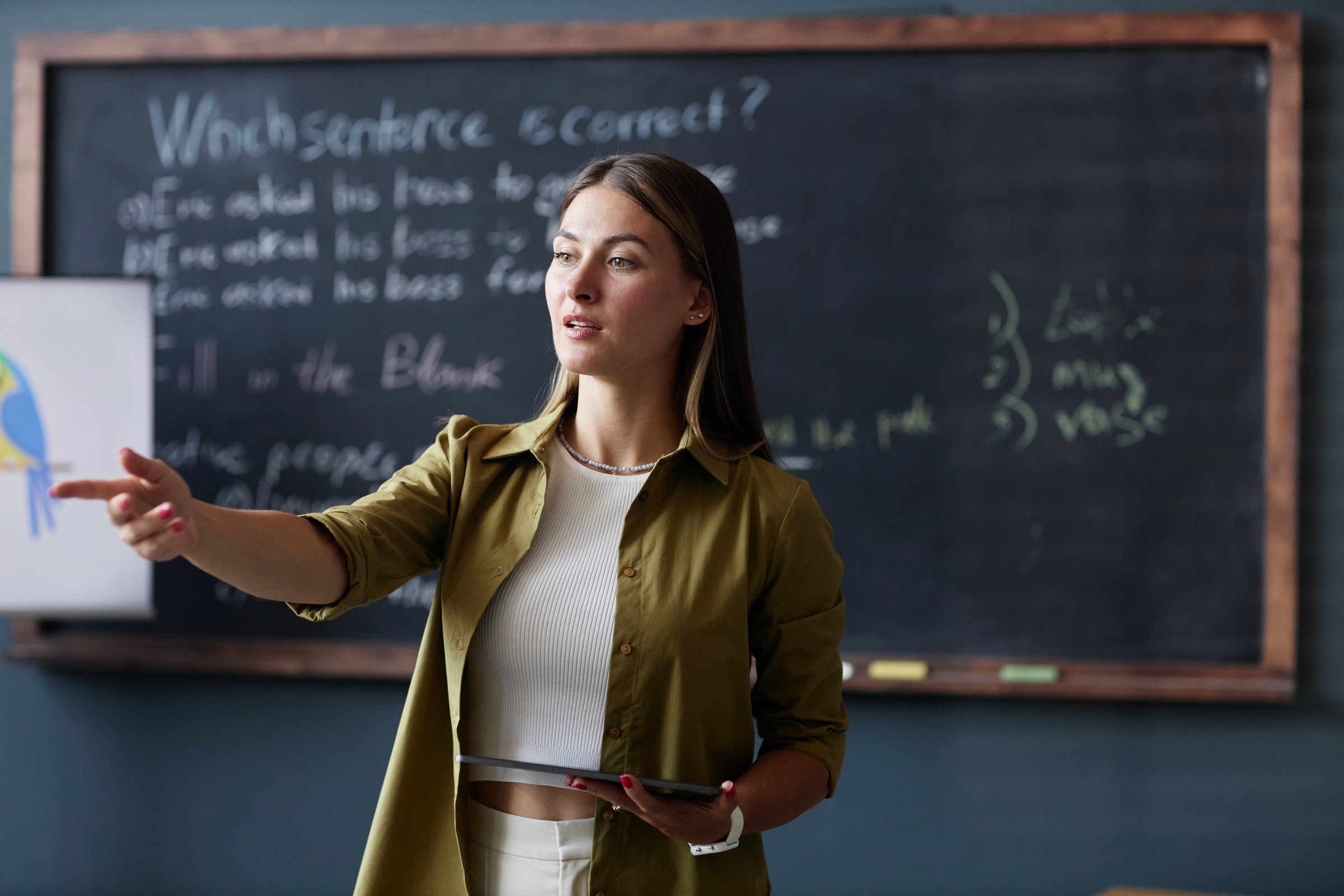Medium shot of female English teacher holding tablet computer addressing student with question, while discussing new grammar rules in group class against blackboard at language school