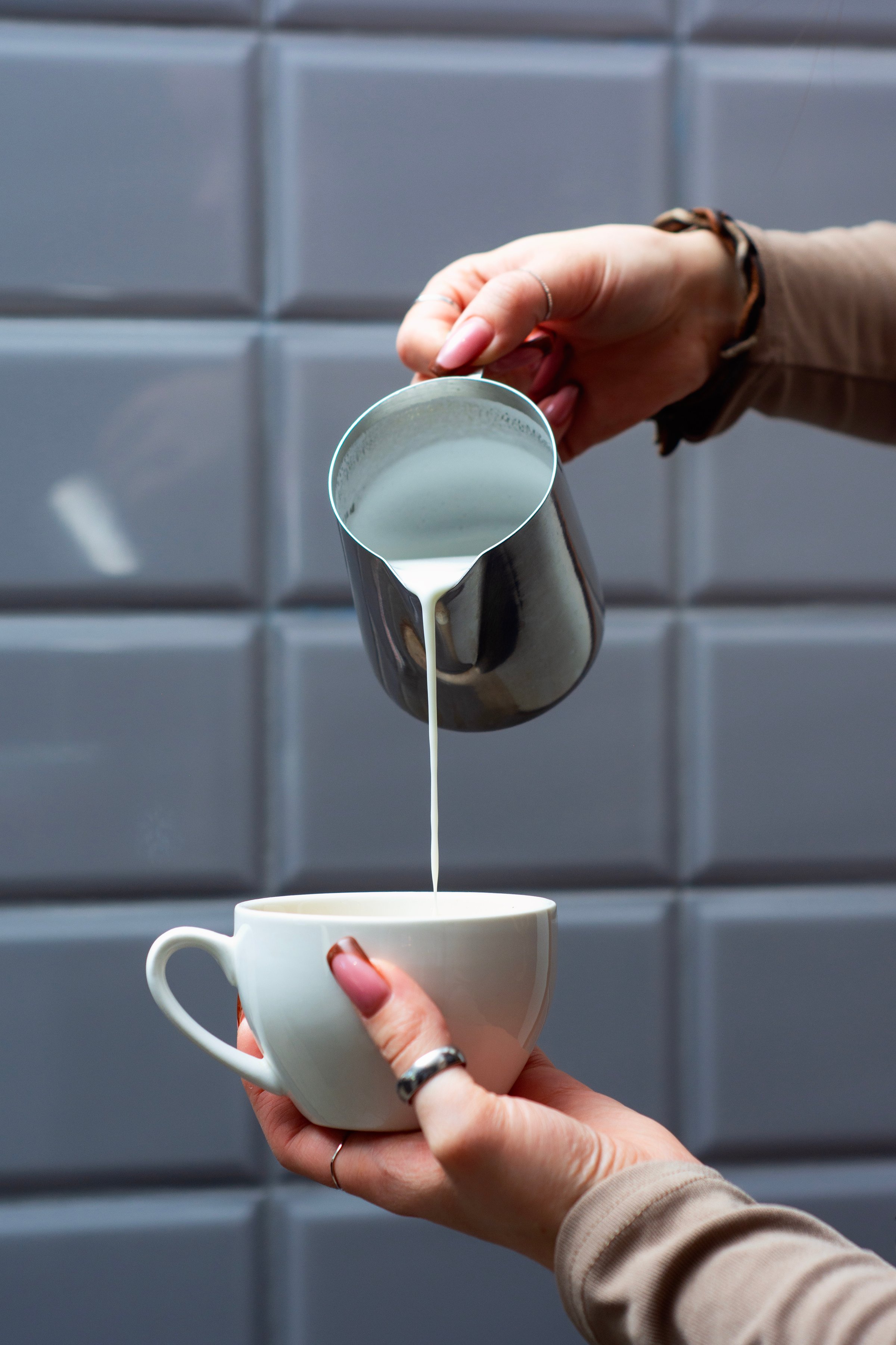 Woman hands pouring creamy milk from a stainless steel pitcher into a white coffee cup on saucer. Professional barista technique for coffee preparation